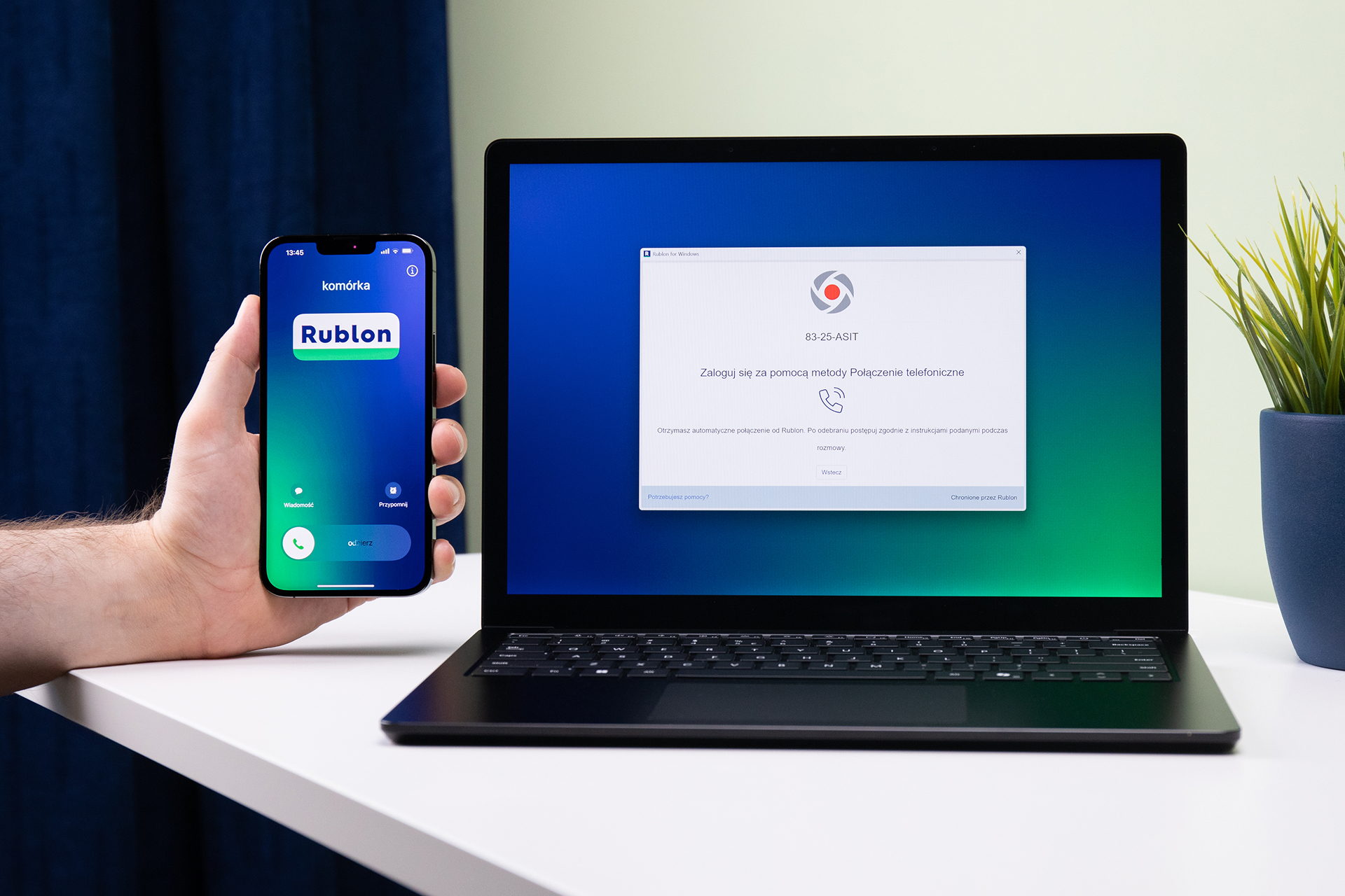 Dłoń trzyma telefon z przychodzącym połączeniem od Rublona; obok laptop z oknem logowania, które czeka na potwierdzenie przez telefon.